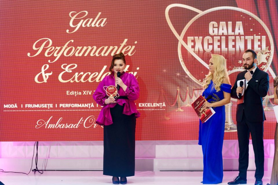Trei persoane stau pe scena Gala Excelenței și a Performanței 2024, cu un fundal roșu și auriu afișând numele evenimentului. O persoană, un talentat designer de rochii de mireasă, deține cu mândrie un premiu.