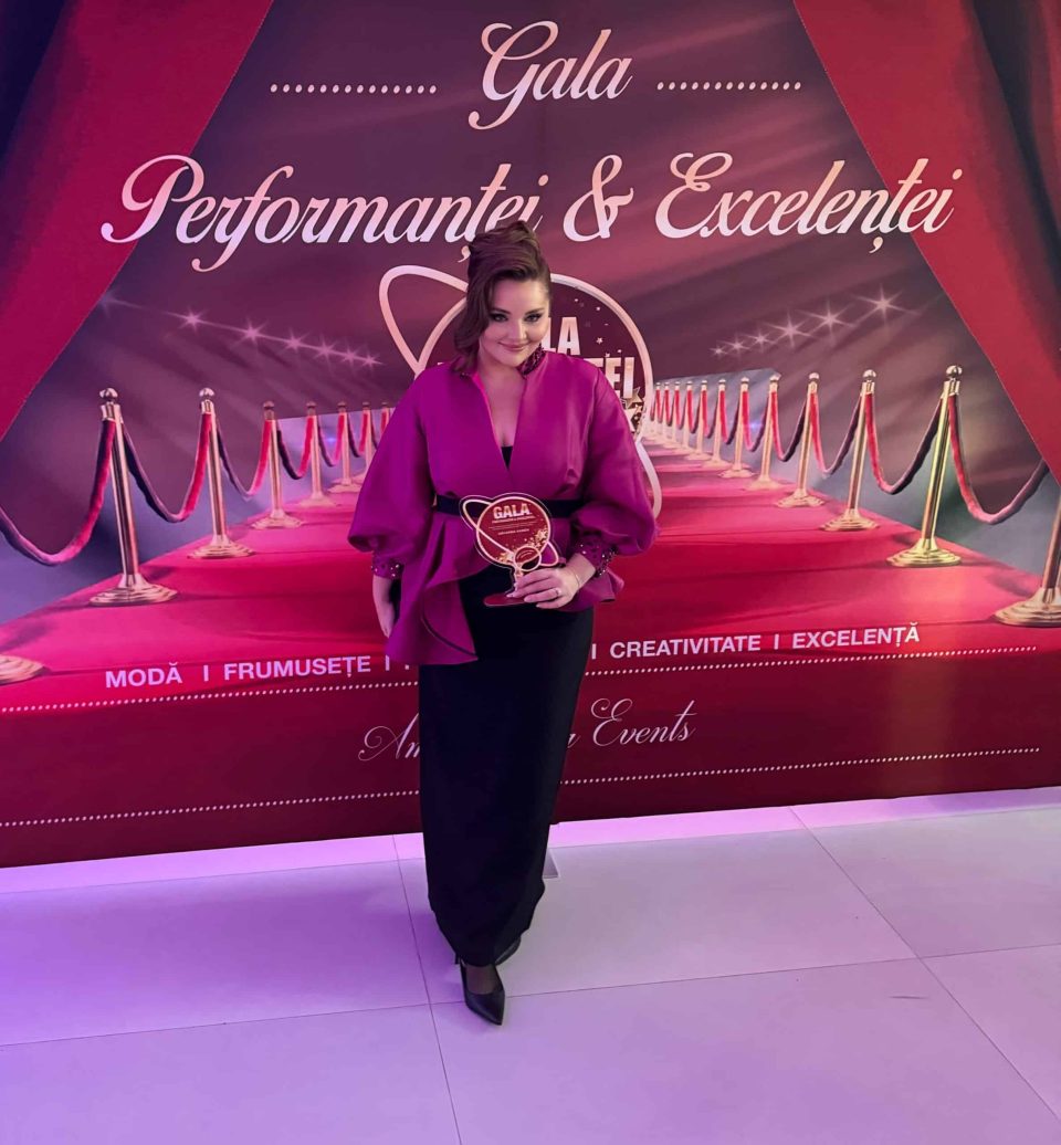 O persoană în ținută mov pozează în fața unui fundal roșu și violet „Gala Excelenței și a Performanței 2024”, ținând cu mândrie un premiu rotund, evidențiind eleganța sinonimă cu România.
