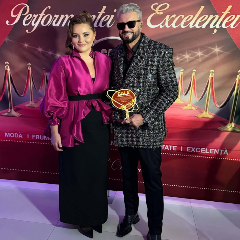 Două persoane care pozează; unul poartă un top roz și o fustă neagră, celălalt o jachetă texturată, ținând în mână un trofeu. Pe fundalul roșu și auriu cu „Performanțe și Excelențe”, acestea strălucesc la Gala Excelenței 2024 din România.