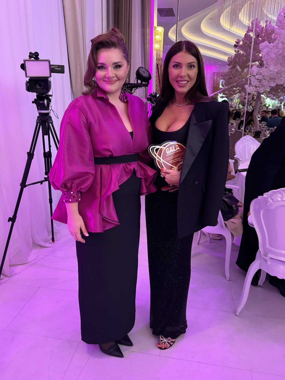 La România Gala Excelenței și a Performanței 2024, două femei pozează. Una strălucește într-o bluză magenta strălucitoare, în timp ce cealaltă uimește într-o rochie neagră cu un blazer. Mesele și luminile decorative pun scena la acest eveniment elegant de interior.
